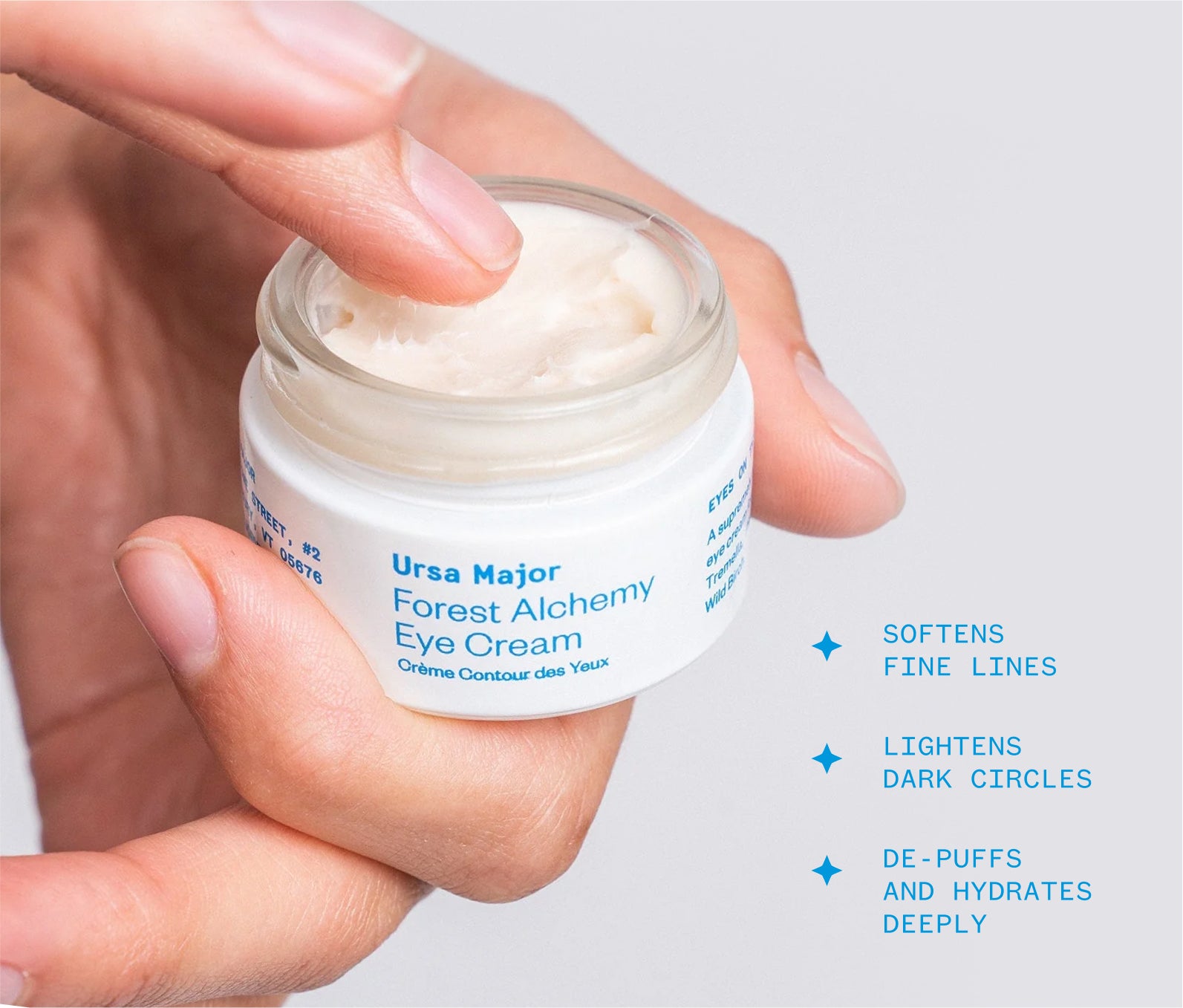 Hand holding an open jar of Ursa Major Forest Alchemy Eye Cream. The image also has text on it that reads this eye cream softens fine lines, lightens dark circle, and de-puffs and hydrates deeply.