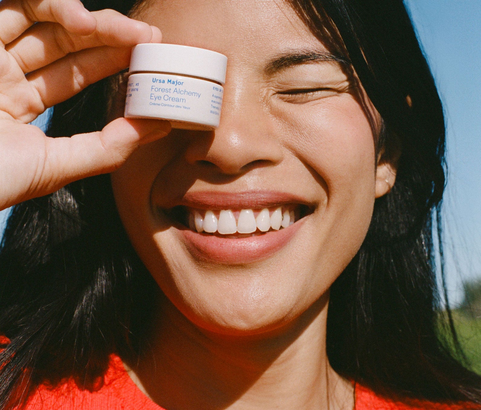 Close-up of a woman holding a jar of Ursa Major Forest Alchemy Eye Cream up to her eye as she smiles into the camera with her eyes closed.