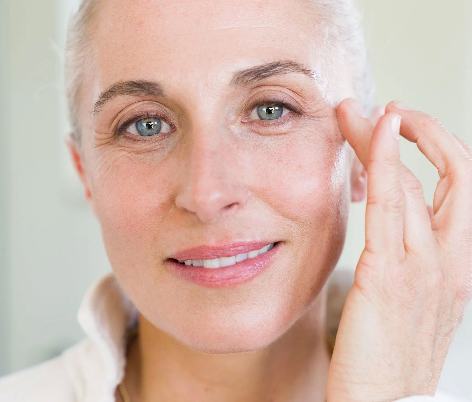 Close-up of a woman looking into the camera slightly smiling.  She is using her ring-finger to swipe Ursa Major Forest Alchemy Eye Cream into her under eye.