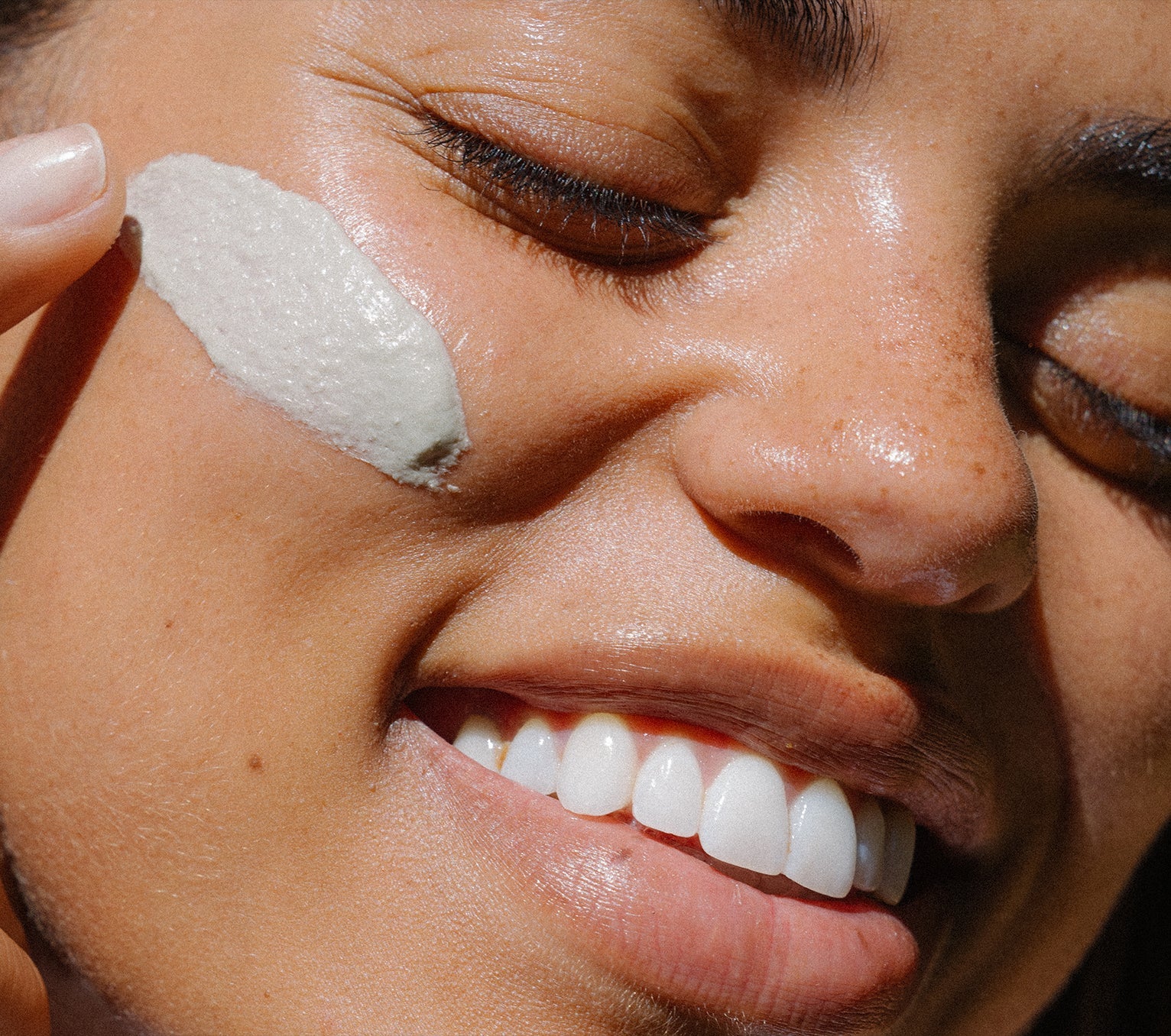 Close-up of a woman's face with Ursa Major Green Slate Mineral Polish swiped on her cheek.  She in smiling with her eyes closed.