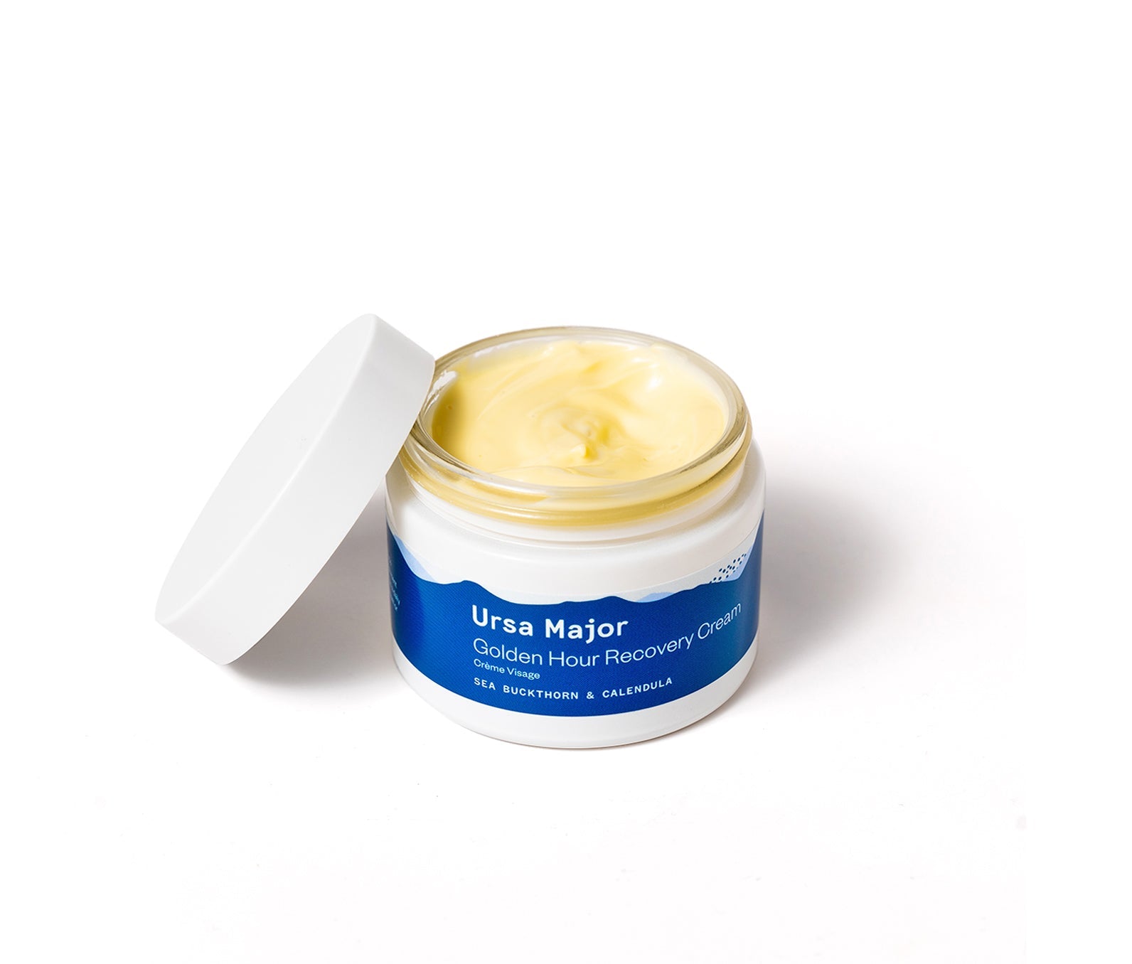 Open jar of Ursa Major Golden Hour Recovery Cream on a white background.  Product in the jar is a light-yellow.  Top of the jar is balanced on the side.