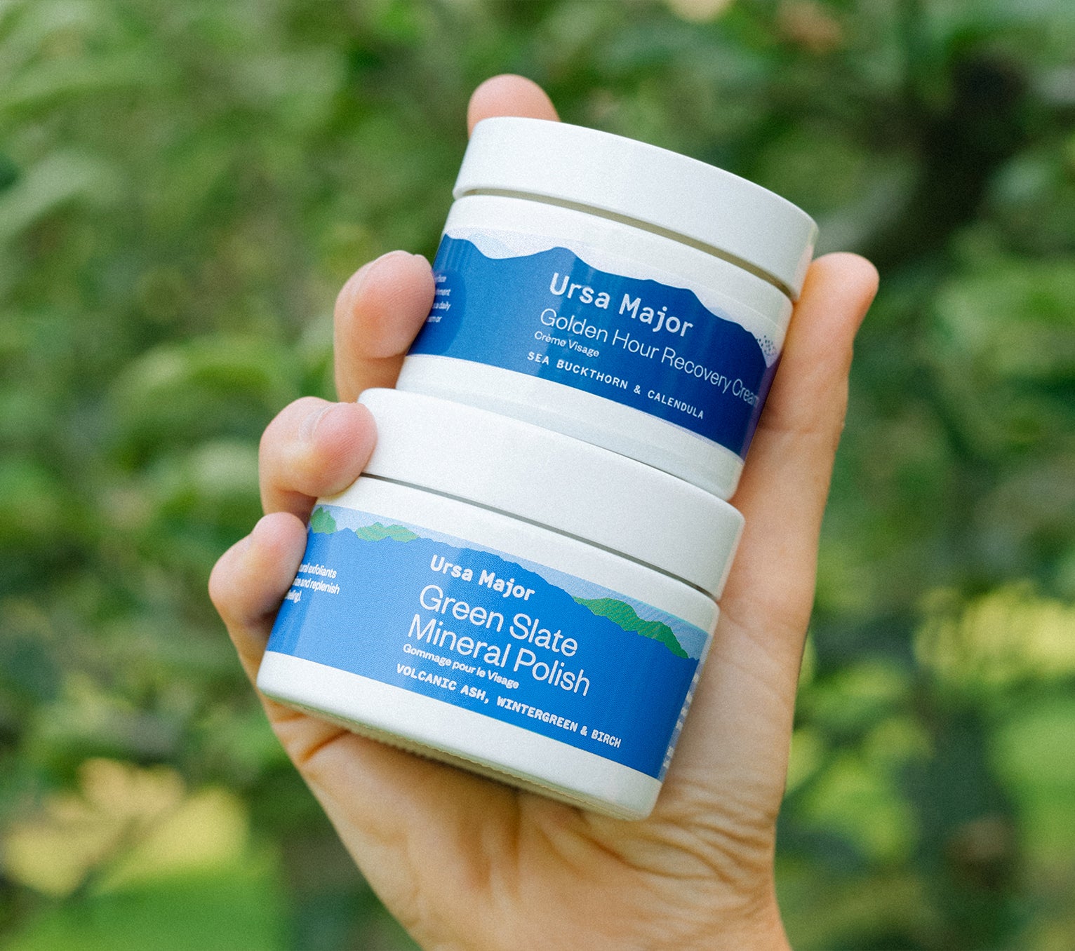 Hand holding a jar of Ursa Major Golden Hour Recovery Cream and Green Slate Mineral Polish outside in front of a green bush.