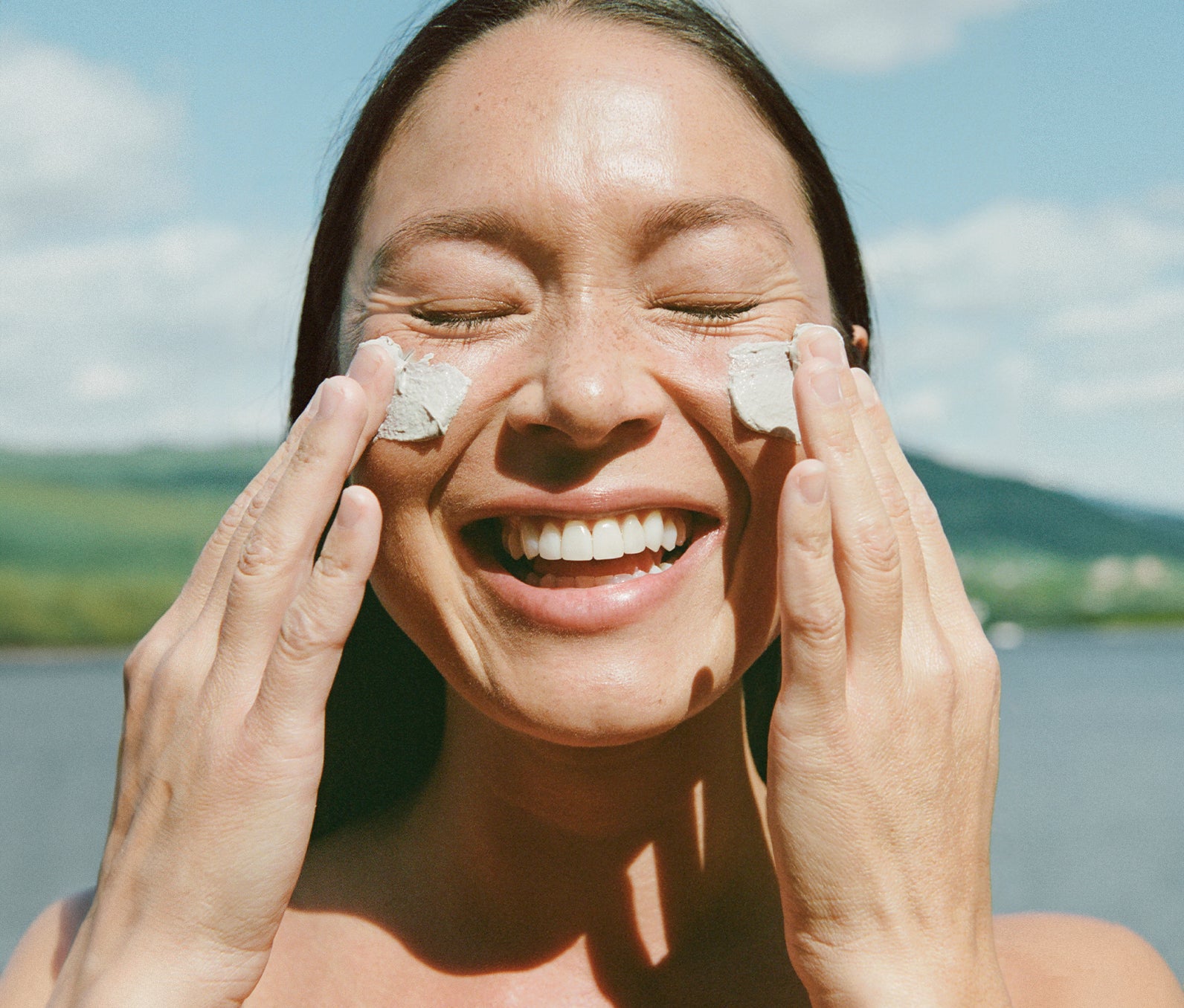 Photo of a woman smiling, standing outside in front of a lake with mountains in the background. She is rubbing Ursa Major Green Slate Mineral Face Polish on her cheeks.