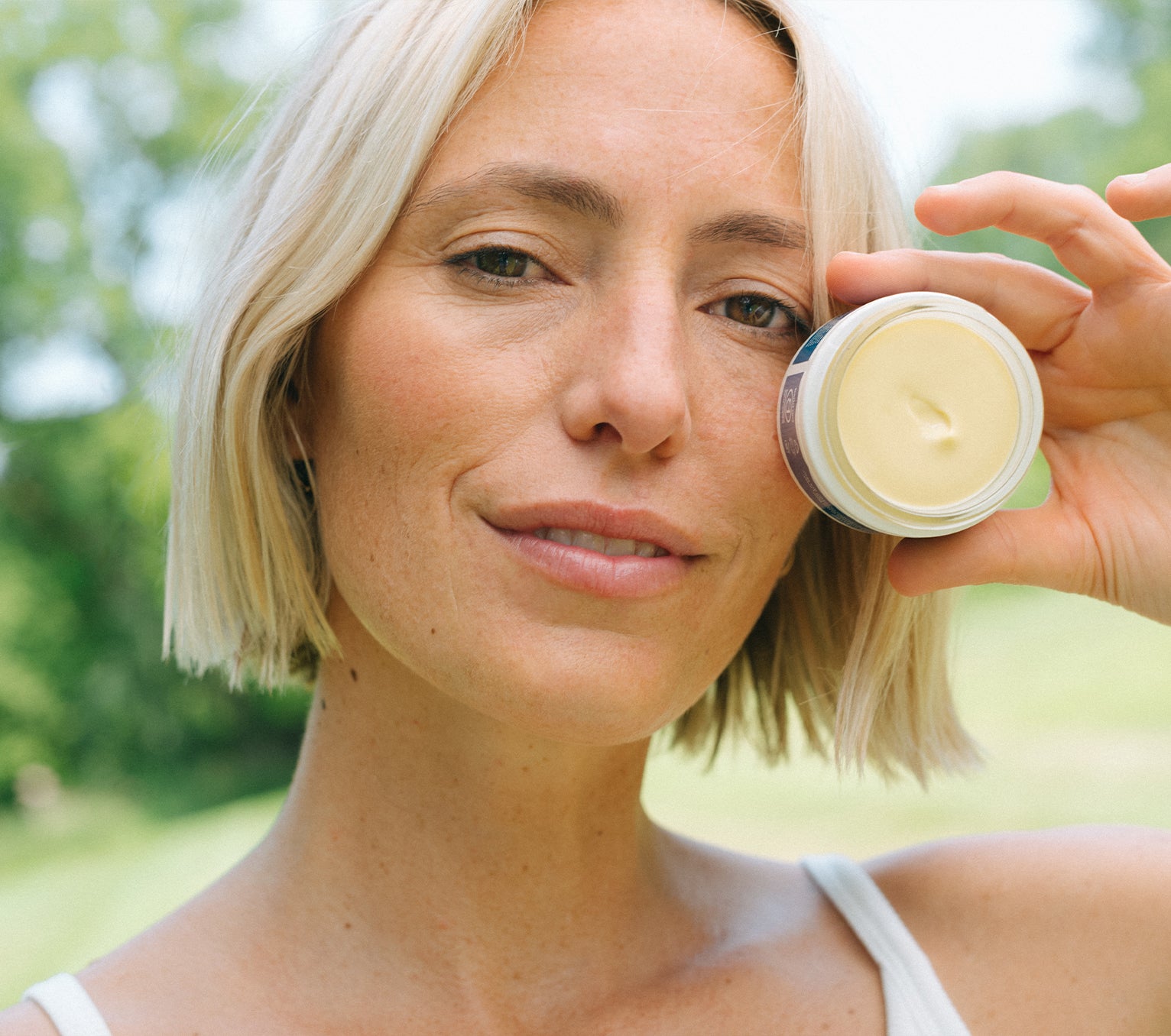 Woman holding an open jar of Ursa Major Golden Hour Recovery Cream outside with trees behind her.