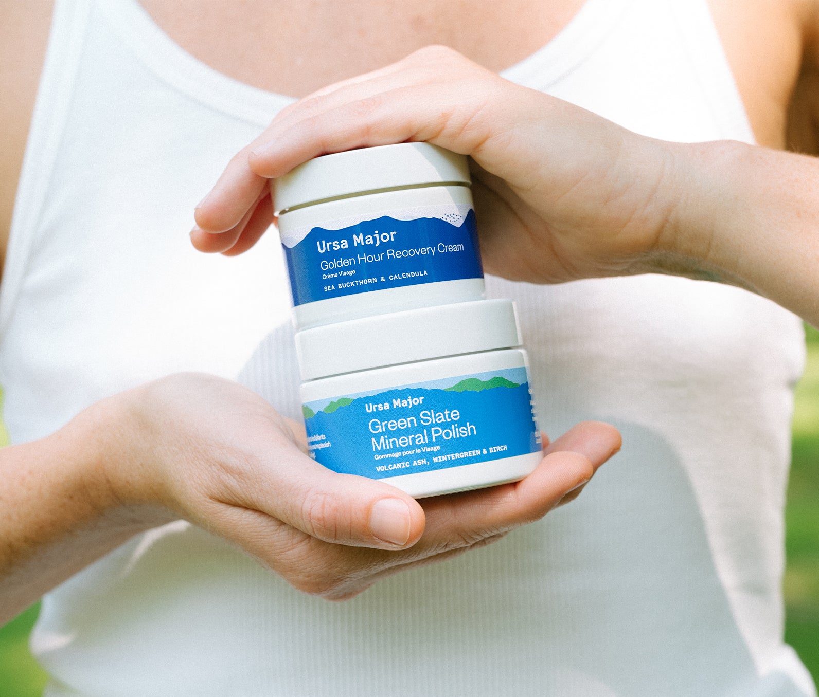 Person holding a jar of Ursa Major Golden Hour Recovery Cream and Green Slate Mineral Polish  between their hands.