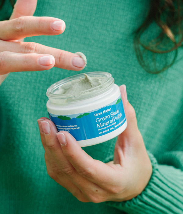 Close up of a person in a green sweater holding an open jar of Ursa Major Green Slate Mineral Polish in one hand and scooping some of the polish out with her finger.