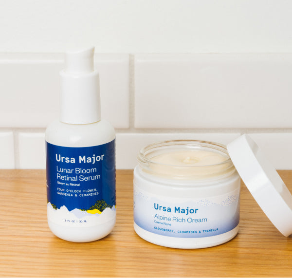 Ursa Major Lunar Bloom Retinal Serum next to an open jar of Alpine Rich Cream on a wooden table with a white tile background.