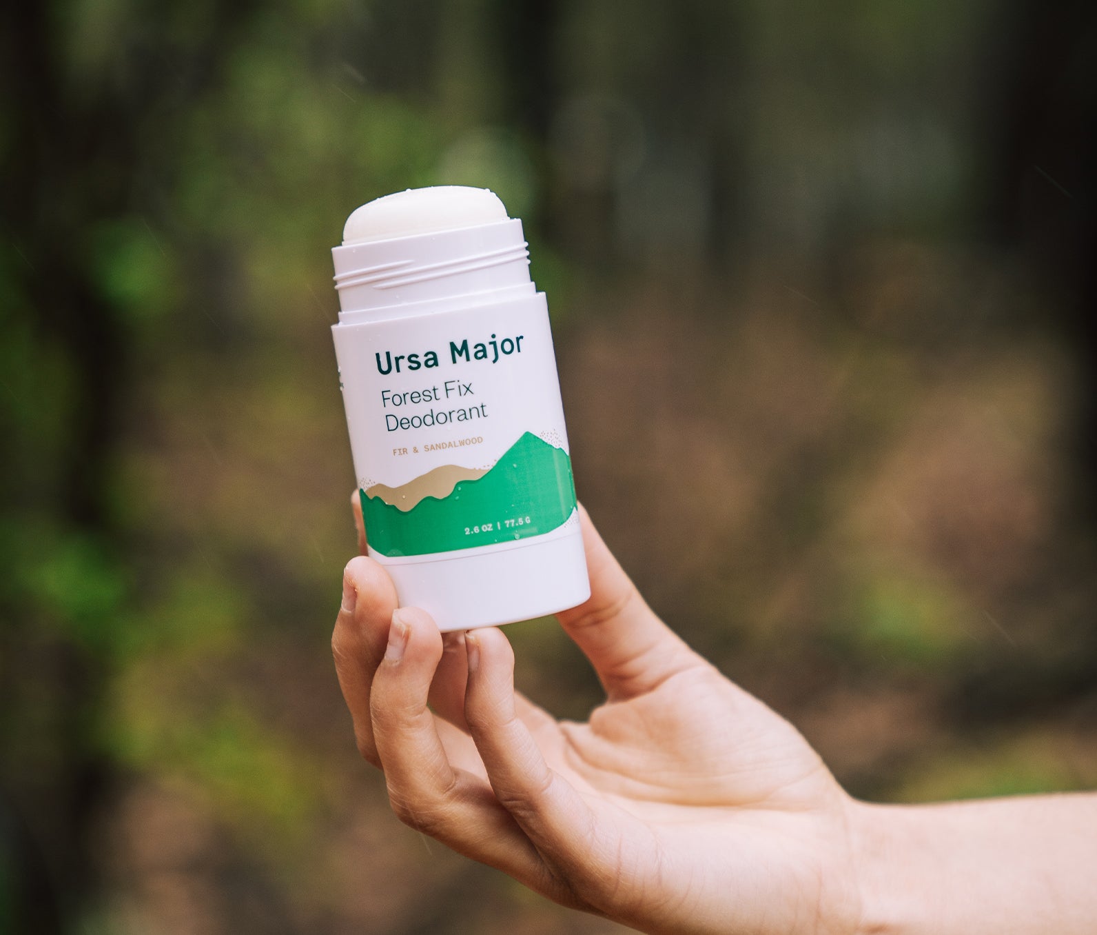 Hand holding an open tube of Ursa Major Forest Fix Deodorant outside in the woods.