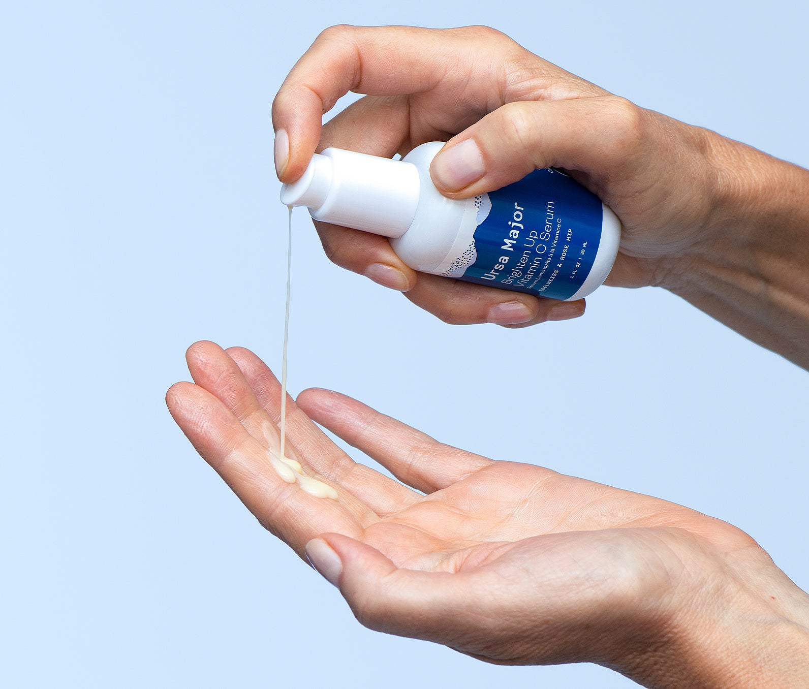 Close-up of hands pumping out Ursa Major Brighten Up Vitamin C Serum from the bottle into their other hand on the light blue background.