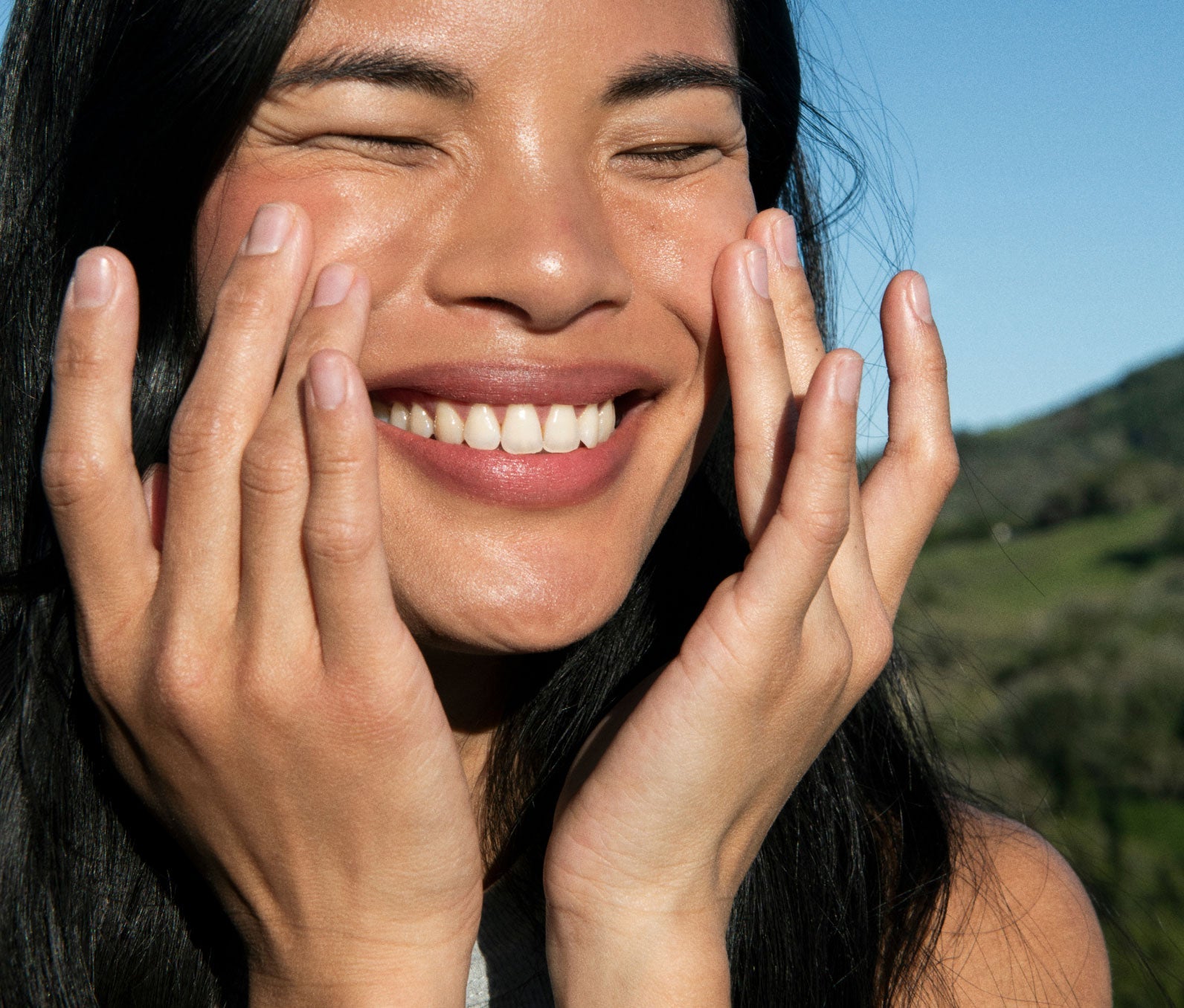 Close-up of a woman's face outside facing the sun.  She is rubbing in Ursa Major Brighten Up Vitamin C Serum onto her face..