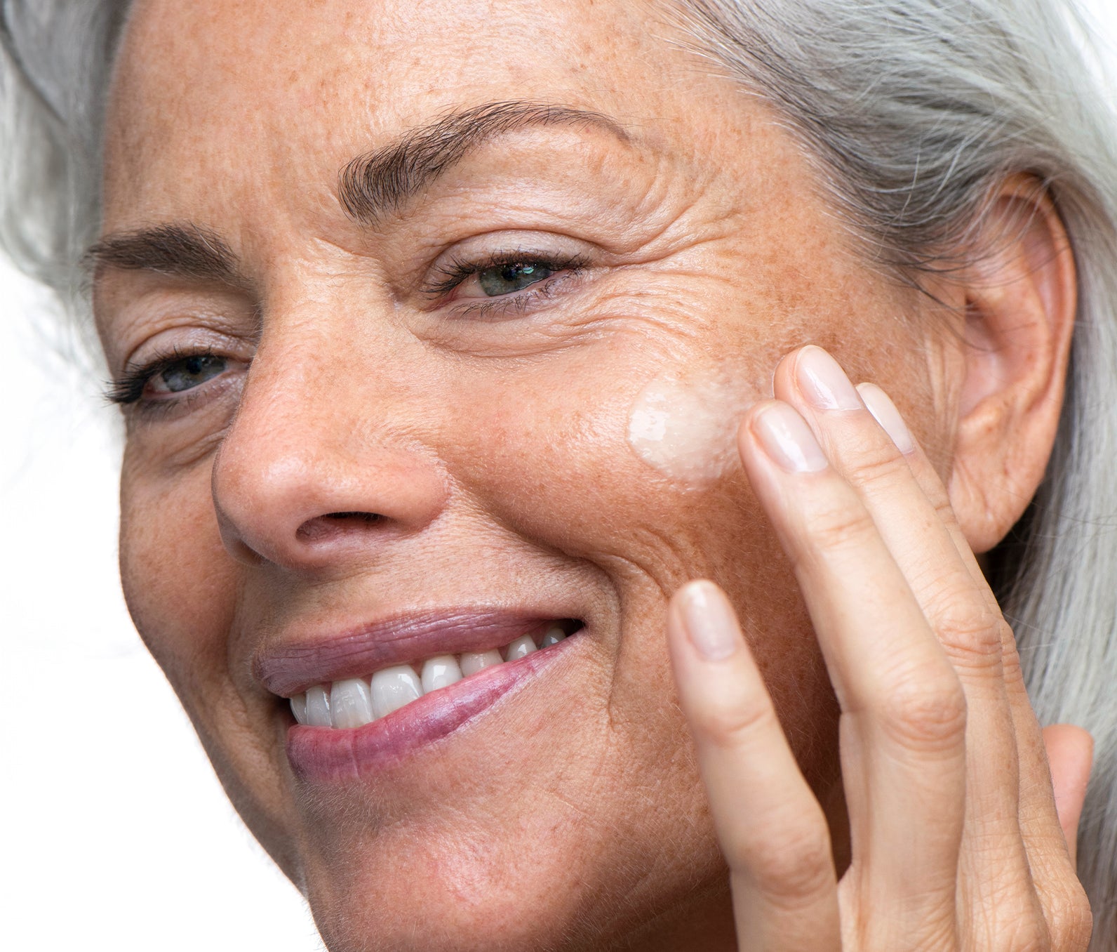 Close-up of an older woman rubbing Ursa Major Brighten Up Vitamin C Serum onto her face with a white background.