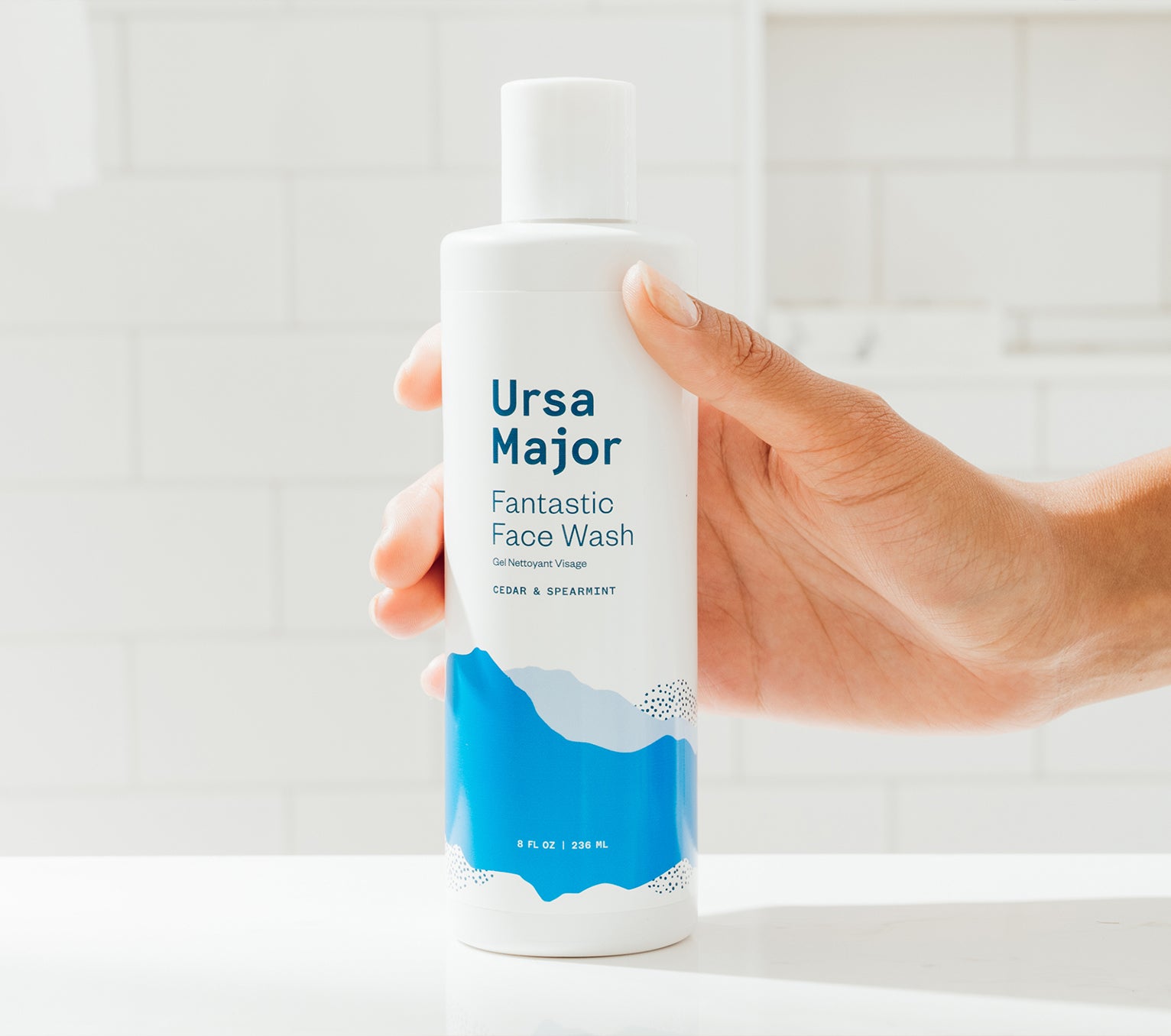 Hand placing a bottle of Ursa Major Fantastic Face Wash onto a white counter against a white tiled wall.