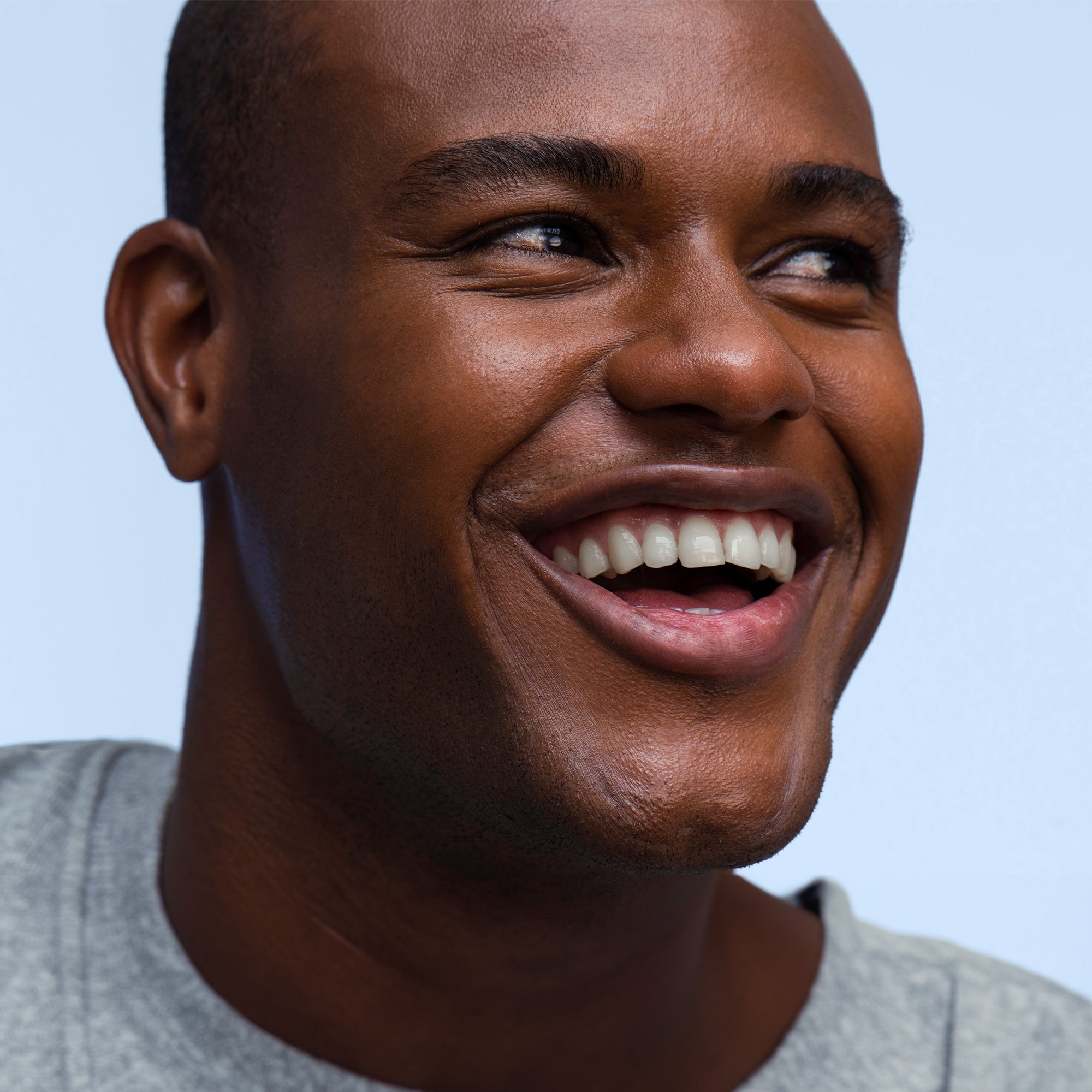 Close-up of a person wearing a gray shirt, smiling looking to the side on a light-blue background.