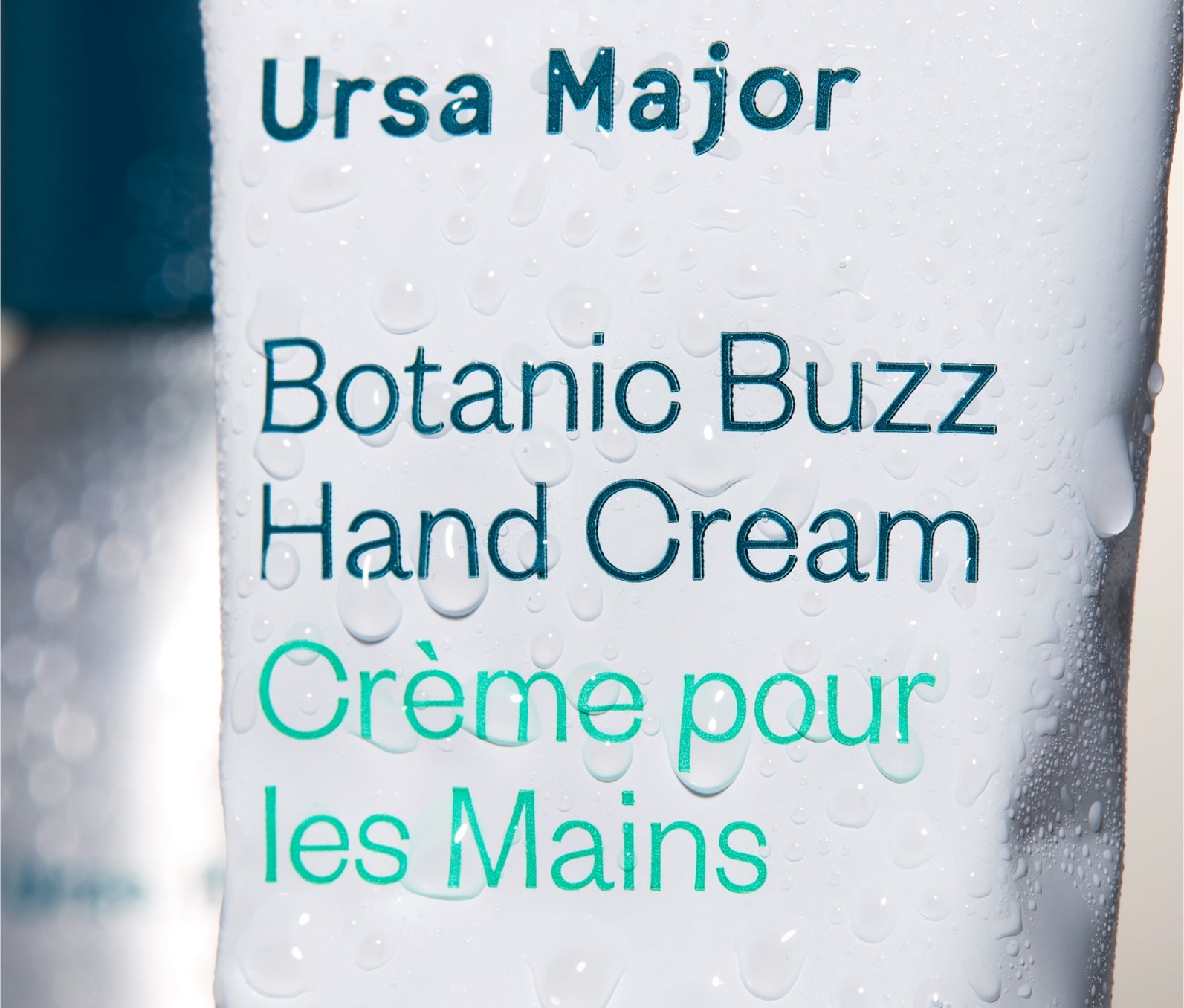 Close-up of the Ursa Major Botanic Buzz Hand Cream tube.  Tube is white with blue lettering.  This image has water droplets on the tube.