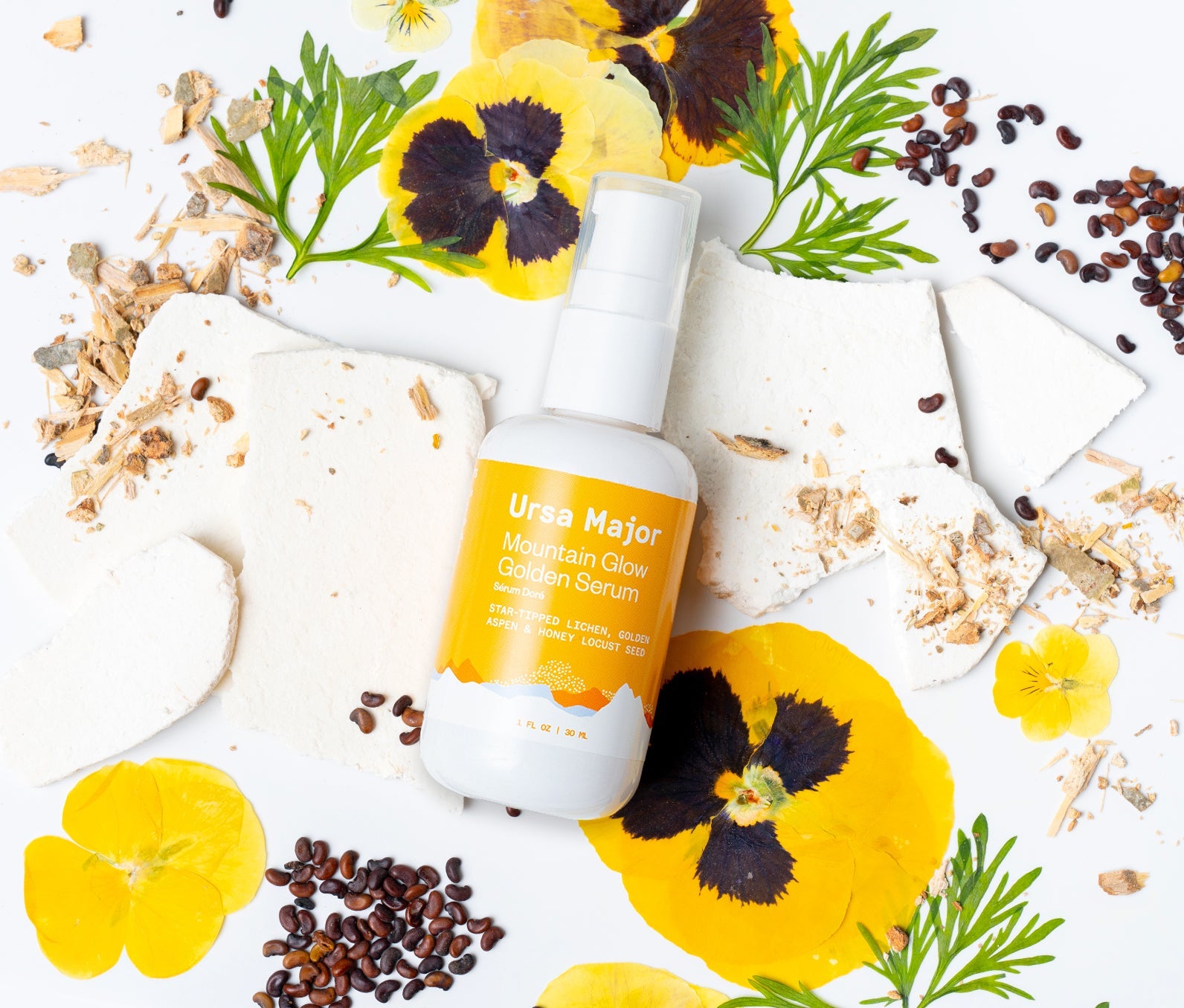 Bottle of Ursa Major Mountain Glow Golden Serum on a white background surrounded by some of the product's natural ingredients. 