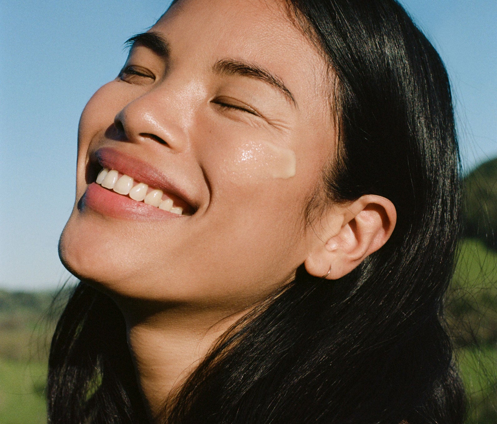 Woman outside smiling with her eyes closed and the sun shining on her face.  She has a swipe of Ursa Major Mountain Glow Golden Serum on her cheek.